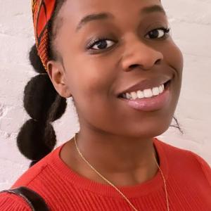 A young Black woman with long hair in a bubble braid, wearing a red and yellow headscarf and a red jumper